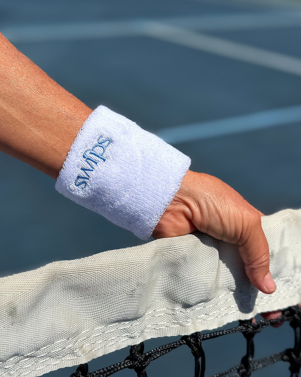 A white Swips wristband worn on a wrist, hand resting on a pickleball net with a blue court behind