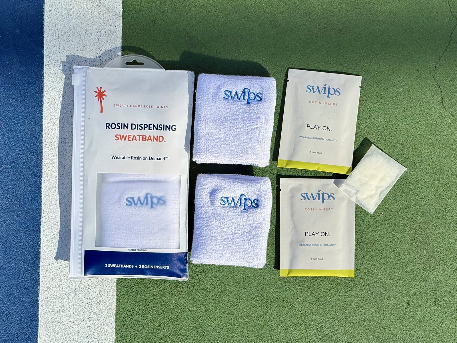 The Swips 2-pack laid out on a pickleball court: retail pouch, two terry cloth sweatbands, two foil sachets and a rosin insert