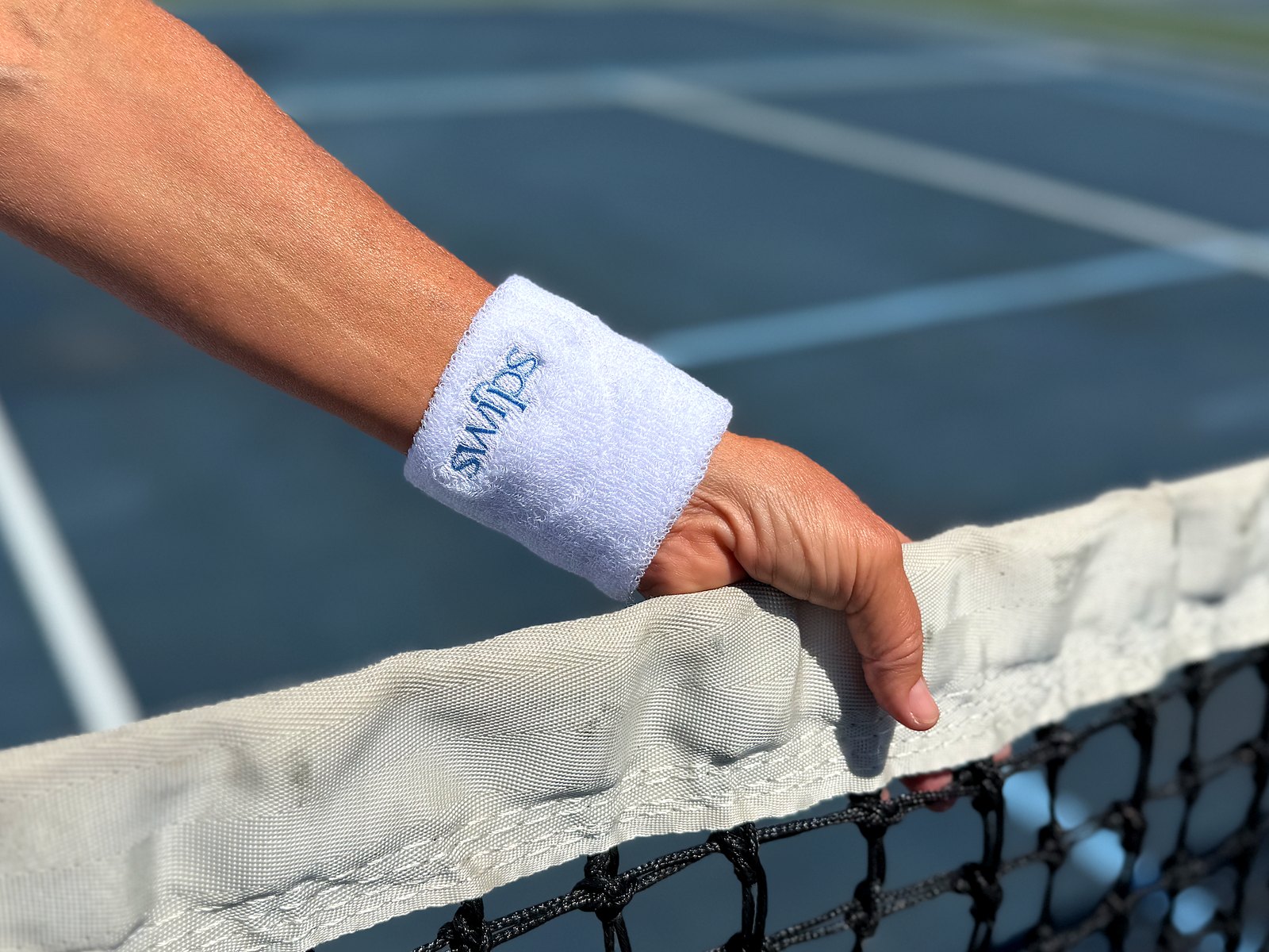 A white Swips wristband worn on a wrist, hand resting on the net of a blue pickleball court