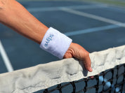 A white Swips wristband worn on a wrist, hand resting on the net of a blue pickleball court